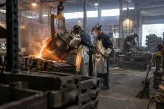Dans une fonderie, deux fondeurs coulent le métal en fusion dans des moules, un jeune ouvrier en veste neuve à leurs côtés