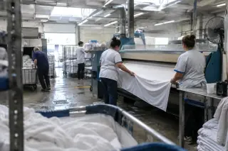 Un jour de chaleur dans une blanchisserie hospitalière : deux employées engagent un drap dans une repasseuse industrielle