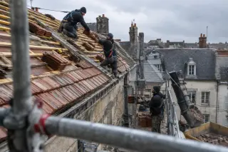 Sur le toit d’une maison en rénovation, deux couvreurs encordés posent des tuiles qu’un collègue monte par l’échafaudage